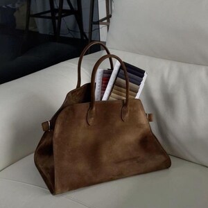 Peut inclure: Un sac fourre-tout en daim marron avec deux poign&eacute;es et une forme rectangulaire. Le sac est ouvert, r&eacute;v&eacute;lant des &eacute;chantillons de tissu de diff&eacute;rentes couleurs. Le sac est pos&eacute; sur une surface blanche.