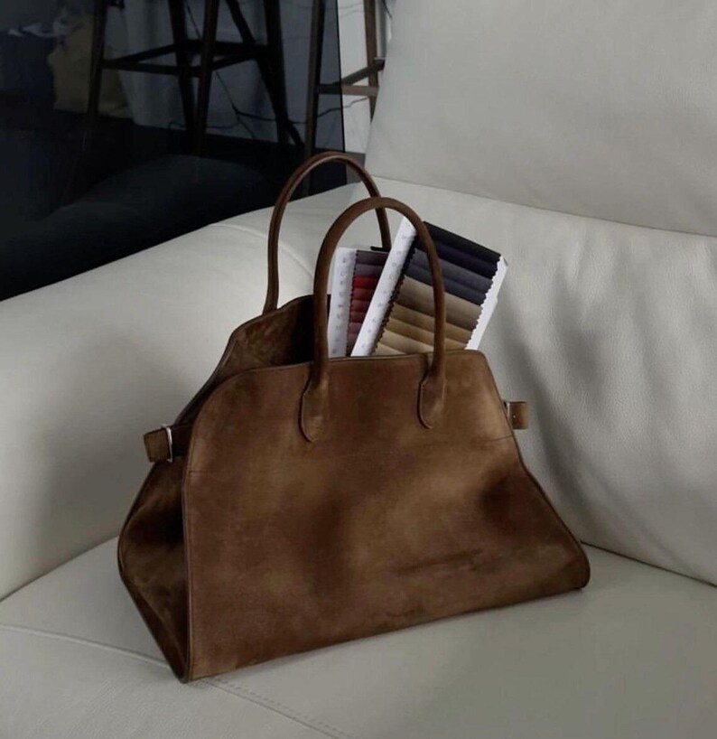 Peut inclure: Un sac fourre-tout en daim marron avec deux poign&eacute;es et une forme rectangulaire. Le sac est ouvert, r&eacute;v&eacute;lant des &eacute;chantillons de tissu de diff&eacute;rentes couleurs. Le sac est pos&eacute; sur une surface blanche.