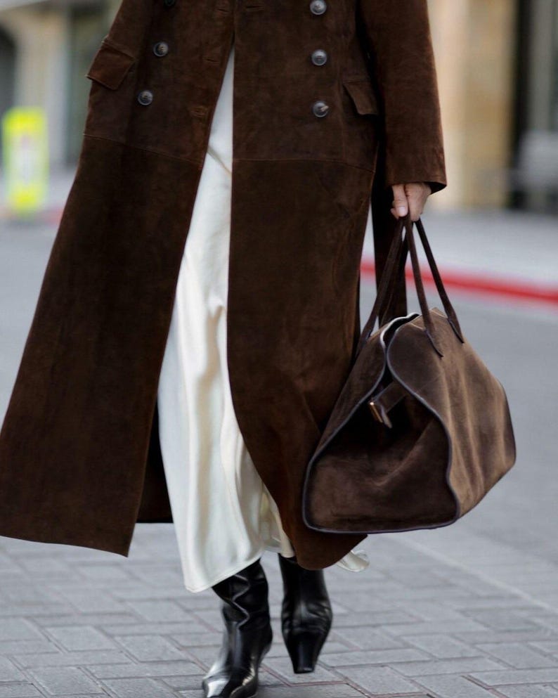 Peut inclure: Un long manteau en daim marron avec de gros boutons et un sac fourre-tout en daim assorti. Le manteau est port&eacute; sur une robe blanche, et la tenue est compl&eacute;t&eacute;e par des bottes en cuir noir.