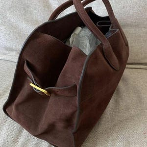 Peut inclure: Un sac fourre-tout en daim marron avec deux poign&eacute;es et une boucle dor&eacute;e. Le sac a une forme rectangulaire avec un int&eacute;rieur spacieux. Le sac est fait d'un mat&eacute;riau doux et textur&eacute;.