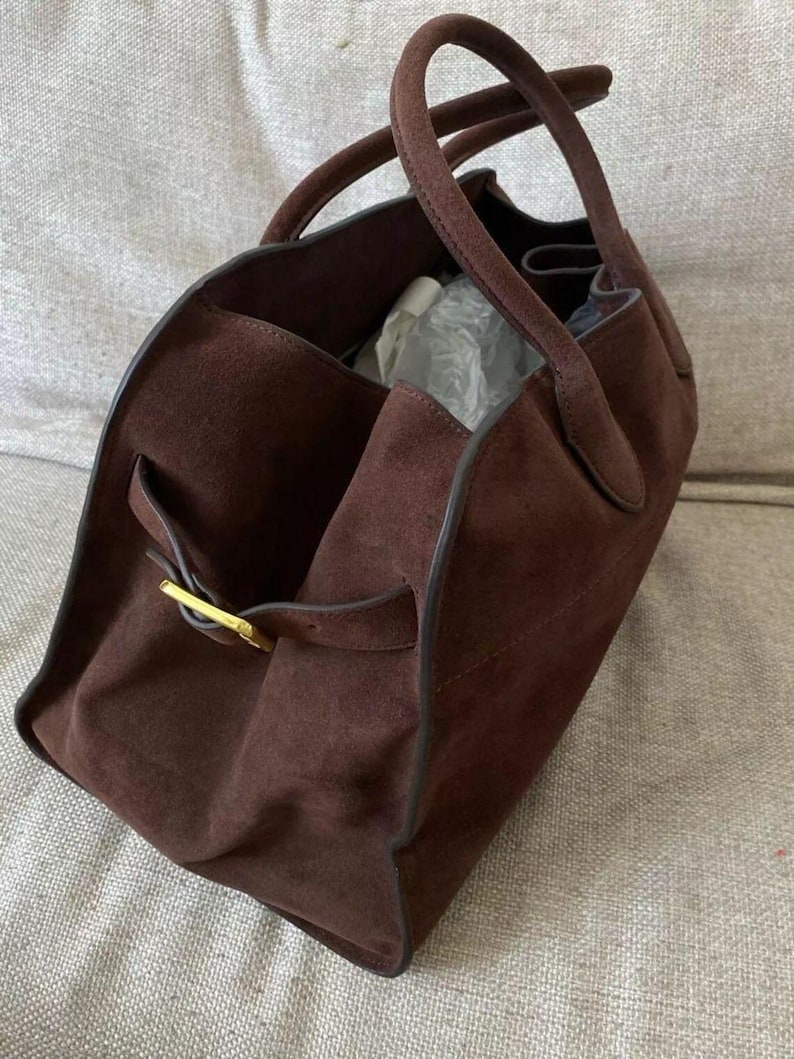 Peut inclure: Un sac fourre-tout en daim marron avec deux poign&eacute;es et une boucle dor&eacute;e. Le sac a une forme rectangulaire avec un int&eacute;rieur spacieux. Le sac est fait d'un mat&eacute;riau doux et textur&eacute;.
