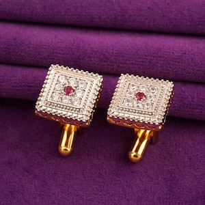 May include: A pair of gold-toned square cufflinks with a central red gemstone surrounded by small clear stones. The cufflinks have a detailed, textured border and a gold post. The background is a deep purple fabric.