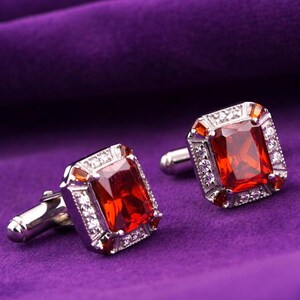 May include: A pair of silver cufflinks with large, rectangular, red gemstones. The gemstones are surrounded by a border of smaller, clear stones. The cufflinks are set against a deep purple background.