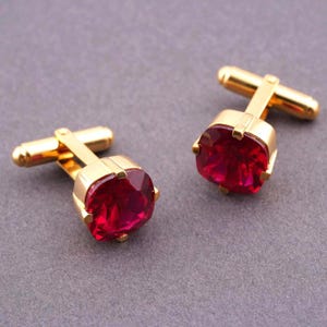 Puede incluir: Un par de gemelos dorados con grandes piedras preciosas cuadradas de color rojo rubí. Los gemelos tienen un diseño clásico, con una barra rectangular y un engaste cuadrado. Las piedras preciosas son de un rojo intenso.