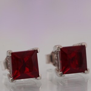 May include: A pair of square-cut, deep red gemstone stud earrings. The gemstones are set in a silver-colored metal with a small, decorative design around the edges. The earrings are displayed against a soft, neutral background.