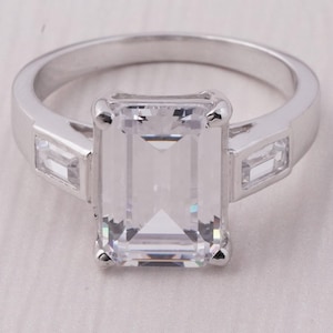 Puede incluir: Un anillo plateado con una gran piedra preciosa rectangular y transparente como punto focal. Dos piedras preciosas rectangulares más pequeñas flanquean la piedra principal. El anillo tiene una banda pulida y un diseño clásico y elegante.