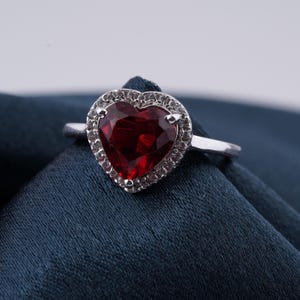 May include: A silver ring featuring a heart-shaped, faceted, deep red gemstone. The heart is surrounded by a halo of smaller, clear stones. The ring is set against a dark blue fabric background, highlighting the ring's sparkle.