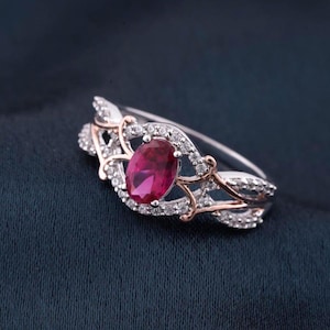 Op de afbeelding: Zilveren ring met een grote ovale robijn in het midden, omringd door kleine, heldere accentstenen. De ring heeft een decoratief ontwerp met roségouden accenten, tegen een donkerblauwe achtergrond.