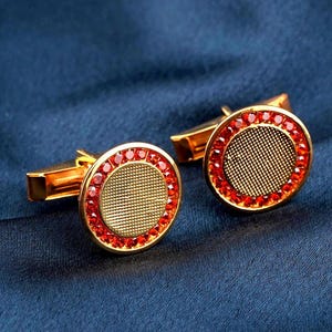 May include: A pair of gold cufflinks with a circular design. Each cufflink has a textured gold centre surrounded by a ring of red gemstones. The cufflinks are displayed on a dark blue fabric.