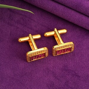 May include: A pair of gold-coloured cufflinks with a rectangular face. The face features small, red gemstones and a border of smaller, clear stones. The cufflinks are displayed against a purple background.