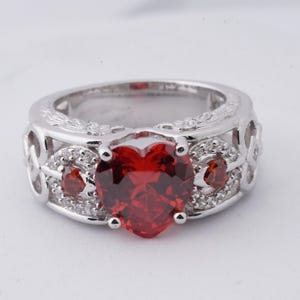 Puede incluir: Anillo plateado con una gran piedra preciosa roja en forma de corazón como pieza central. El anillo tiene piedras preciosas rojas más pequeñas y piedras de acento transparentes. La banda presenta un diseño calado con detalles decorativos.