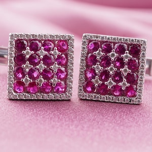 May include: A pair of square cufflinks with a silver frame. Each cufflink features a central arrangement of round, deep pink gemstones, with smaller, clear stones. The cufflinks are set against a pink background.
