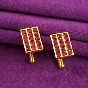 May include: A pair of gold-toned cufflinks with a rectangular design. Each cufflink features a grid of small, square, red gemstones. The cufflinks are displayed on a purple fabric background.