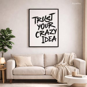 May include: A black framed print with the words "TRUST YOUR CRAZY IDEA" in black brushstrokes. The print hangs above a beige sofa with cushions and a throw. A small side table with a lamp and a plant are to the left.