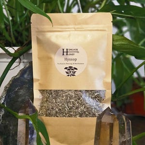 May include: A brown paper bag with a clear window, filled with dried herbs. The bag has a circular label with the text "Hemlock Honey Hyssop" and a botanical illustration. Two clear quartz crystals flank the bag.