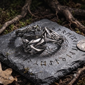 Op de afbeelding: Drie zilverkleurige metalen ringen met een textuur van doornige ranken. De ringen zijn gerangschikt op een donkergrijze, vierkante stenen plaat die is gegraveerd met cirkelvormige patronen en runentekens, met een kleine metalen munt aan de rand.