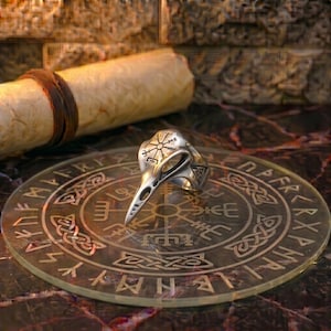 May include: A silver-toned metal ring shaped like a raven skull, featuring an engraved Vegvisir symbol on the forehead. The ring rests on a circular glass plate etched with runic symbols and a central compass design, with a rolled parchment scroll nearby.