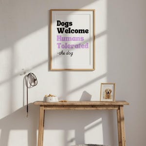 May include: A framed print with the text "Dogs Welcome Humans Tolerated -the dog" hangs above a wooden table. A dog bed, a dog bowl, and a framed dog photo are on the table. A dog leash hangs on the wall.