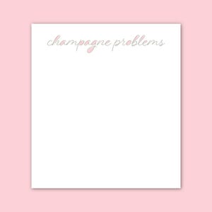 May include: A white notepad with the phrase "champagne problems" in a cursive font, with pink accents, against a pink background. The notepad is rectangular and has a clean, minimalist design.