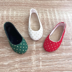 Peut inclure: Trois ballerines miniatures vertes, blanches et rouges, chacune ornée de pois dorés. Les chaussures sont disposées sur une surface en bois brun clair, mettant en valeur leur design délicat et leurs couleurs vives.