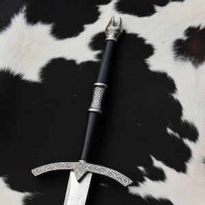 Könnte beinhalten: Ein mittelalterliches Schwert mit einem schwarzen Griff und einem silberfarbenen Parierstange und Knauf. Die Klinge ist silberfarben und der Griff hat dekorative silberne Bänder. Das Schwert wird vor einem schwarz-weißen Kuhfellhintergrund präsentiert.