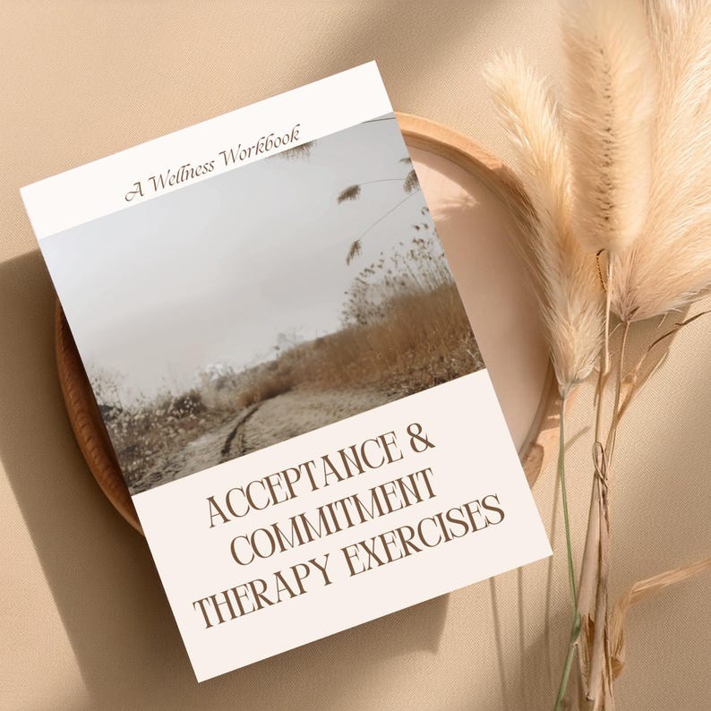 May include: A wellness workbook titled "Acceptance & Commitment Therapy Exercises" with a landscape image on the cover. The book is set on a wooden tray with dried ornamental grass in the background. The color palette is neutral with beige and brown tones.