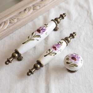 Op de afbeelding: Twee langwerpige keramische ladehandgrepen en één ronde keramische knop, alle met een witte basis en handgeschilderde paarse bloemen en groene bladeren. Elk stuk is voorzien van antiek messingkleurig metalen beslag aan de uiteinden.