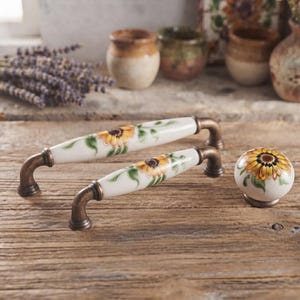 May include: Two ceramic drawer pulls and one matching round cabinet knob, each featuring a hand-painted yellow sunflower design with green leaves on a white background, finished with antique bronze metal hardware on the ends.