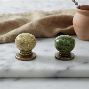 Può includere: Due pomelli rotondi per mobili con finitura lucida, uno in verde oliva chiaro screziato e l'altro in verde bosco intenso. Ogni pomello presenta una parte superiore sferica liscia montata su una base metallica circolare color ottone antico.