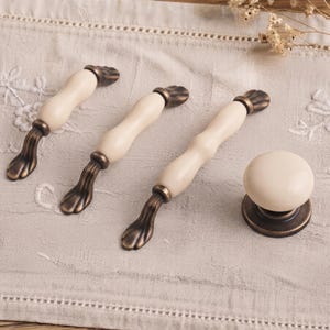 May include: Three elongated cabinet pulls and one round cabinet knob. Each piece features a smooth, cream-colored ceramic center section paired with an antique bronze-finished metal base and ends, showcasing a detailed, textured design.