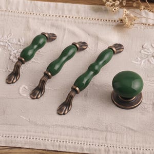 May include: Three elongated cabinet pulls and one round cabinet knob, all featuring a glossy dark green ceramic center with antique bronze-toned metal ends. The pulls have a wavy, organic shape, while the knob is a smooth, circular design.