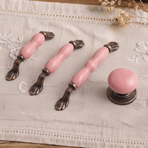 May include: Three pink ceramic drawer pulls with antique bronze metal bases and one matching round pink ceramic knob with an antique bronze base. The pulls feature a curved, elongated shape with metallic accents at the ends and center.