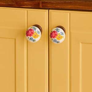 May include: Two floral ceramic cabinet knobs with brass fittings. The knobs have a white base with a pattern of pink roses, yellow flowers, and blue accents. They are mounted on a yellow cabinet.
