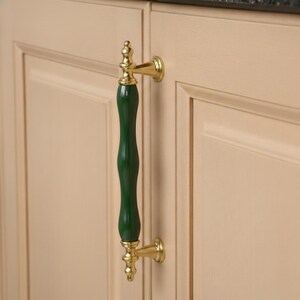 Peut inclure: Une poignée de meuble avec un centre allongé vert foncé et des accents dorés. La poignée est fixée à une porte d'armoire beige clair. La quincaillerie a un design classique et décoratif.