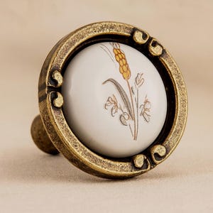 Könnte beinhalten: Antiker Messing-Schrankknauf mit weißem Keramikmittelteil. Der Keramikeinsatz zeigt ein Weizenähren-Design in Gelb- und Brauntönen. Der Knauf hat einen dekorativen, verzierten Rahmen.