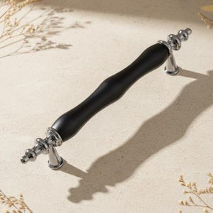 May include: A cabinet pull with a black, curved center and silver-colored, decorative ends. The hardware is designed for furniture and has a modern aesthetic. The pull is shown on a neutral background with shadows.