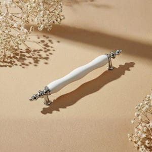 Può includere: Una maniglia per mobili bianca e argentata con un centro curvo in ceramica bianca e estremità argentate ornate. L'hardware è ambientato su uno sfondo neutro con delicati fiori bianchi, suggerendo un tema di arredamento per la casa.