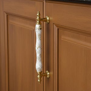 May include: A cabinet pull with a white and gold speckled handle and gold-toned hardware. The decorative handle is mounted on a brown cabinet door. The pull is designed for easy opening and closing of cabinet doors.