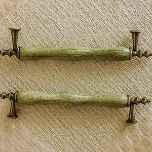 May include: Two cabinet pulls. Each has a green, textured central section and bronze-coloured hardware. The pulls have a cylindrical, slightly curved design with decorative end pieces. The green centre has a marbled appearance.