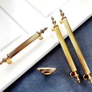 May include: Gold-toned cabinet hardware set. The set includes two long, ribbed handles, a shorter handle, and a smaller knob. The hardware has ornate detailing on the ends and a polished finish. The hardware is designed for cabinets and drawers.