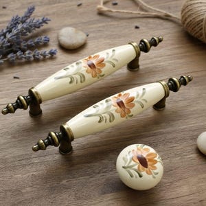 Op de afbeelding: Crèmekleurige kastgrepen en een knop met een bloemmotief in oranje en groen. De hardware heeft bronzen metalen accenten. De grepen zijn ongeveer 15 cm lang. De knop is rond en heeft een vergelijkbaar bloemmotief.