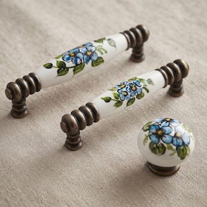 May include: Two elongated ceramic drawer pulls and one round ceramic knob, each featuring a white base with hand-painted blue flowers and green leaves. The hardware includes antique bronze-toned metal ends with a ribbed, circular design.