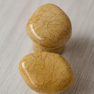 Pode incluir: Dois puxadores de armário ovais e lisos, com um tom castanho-amarelado quente e padrões de veios intrincados. Os puxadores são exibidos contra um fundo claro e texturizado, sugerindo uma aparência de pedra natural ou mármore.