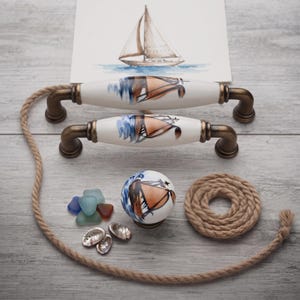 Può includere: Due maniglie per cassetti in ceramica e un pomello rotondo con un disegno di barca a vela dipinto e ferramenta in metallo color bronzo. Il set include un pezzo di corda arrotolata, diversi piccoli pezzi di vetro marino colorato e tre conchiglie.