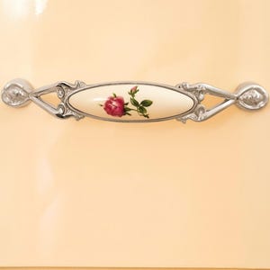 May include: Cream-colored cabinet door with a decorative handle. The handle features a central oval porcelain insert with a pink rose and green leaves. The handle's metal frame is silver-toned with ornate detailing.