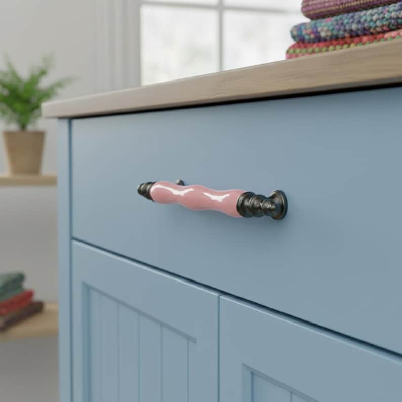 May include: A pale blue cabinet with a wooden top and a pink and bronze handle. The handle is a wavy shape with bronze ends. The cabinet has a paneled door design.
