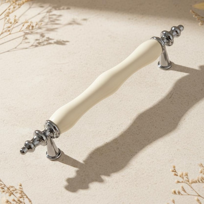 May include: Cream-coloured cabinet pull with a curved, ivory-coloured centre and polished silver-tone metal accents. The hardware features decorative detailing on the ends and a classic design. Suitable for kitchen or bathroom cabinets.