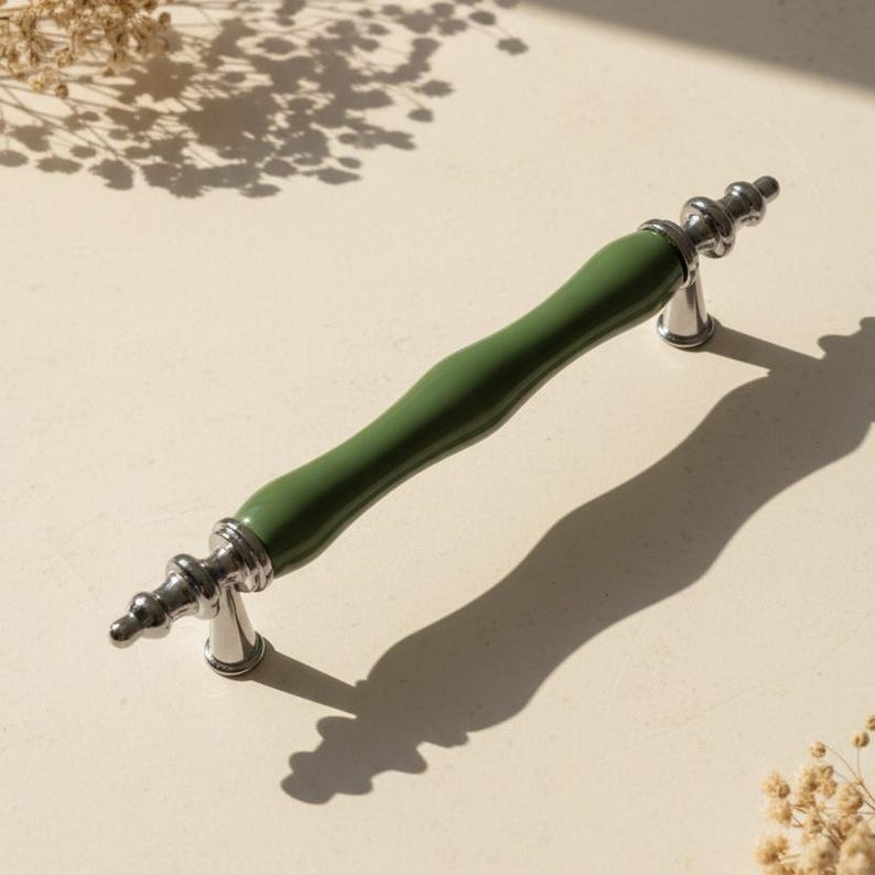 May include: A green and silver cabinet pull handle. The handle has a curved, olive green centre section and silver-coloured, decorative end pieces. The handle is approximately 15 cm long.