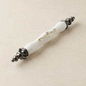 Pode incluir: Puxador de armário branco e dourado com detalhes em preto. O centro do puxador é branco com um design decorativo dourado. As extremidades são pretas com um design arredondado. Esta ferragem de armário é um detalhe decorativo para móveis.