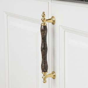 Pode incluir: Um puxador de armário com acabamento dourado e centro castanho escuro, semelhante a mármore. O puxador está preso a uma porta de armário branca. O acabamento dourado tem um design decorativo em cada extremidade.